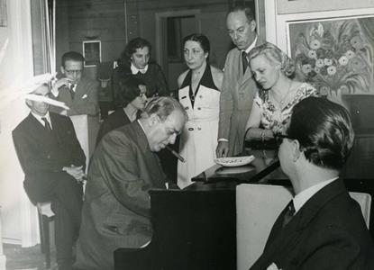 Heitor Villa-Lobos ao piano, interpretando “A lenda do caboclo”. Léonide Massine (sentado, à esquerda), Fructuoso Vianna (primeiro, de pé, à esquerda), Annette Celine, Felicja Blumental (de branco, ao centro) e Julieta Strutt (irmã de Mindinha Villa-Lobos, escorada ao piano). Os demais sem identificação. (Foto: Acervo de Annette Celina).